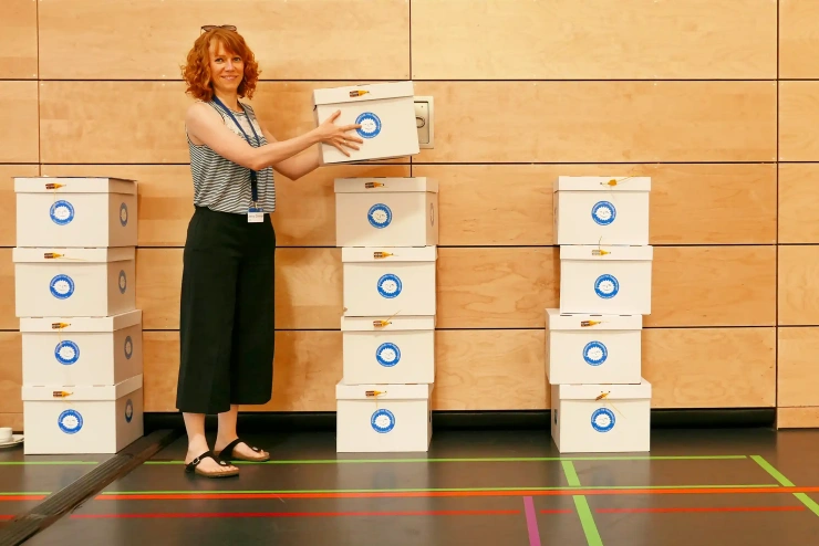 Vor einer Holzwand hebt eine junge, sommerlich gekleidete Frau einen Karton auf einen Stapel weiterer Kartons.