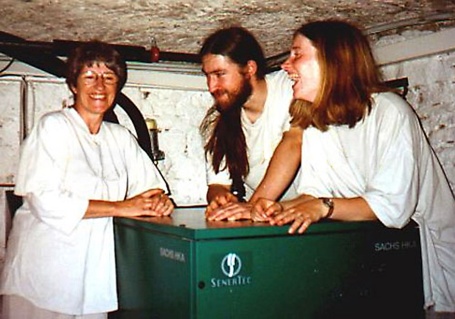 Drei fröhliche Menschen lehnen in einem niedrigen Keller an einem hüfthohen Blockheizkraftwerk.