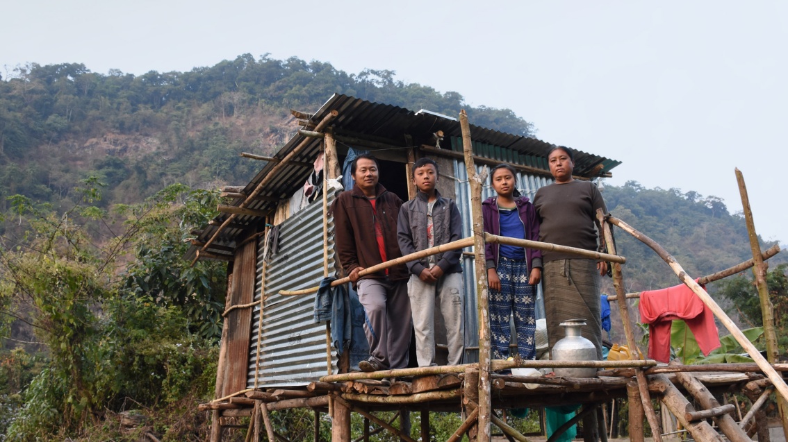 Eine Familie mit zwei Kindern steht auf der Plattform einer Wellblechhütte in Stelzenbauweise, vor ihnen ein Wasserbehäter.