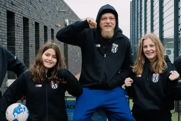 Fünf Junge Menschen in Vereinskleidung stehen bei einem Stadion und lachen in die. Kamera.