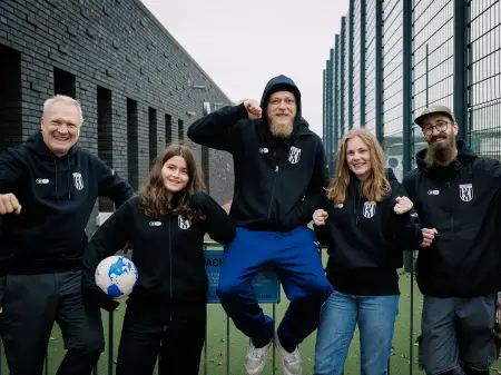 Fünf Junge Menschen in Vereinskleidung stehen bei einem Stadion und lachen in die Kamera.