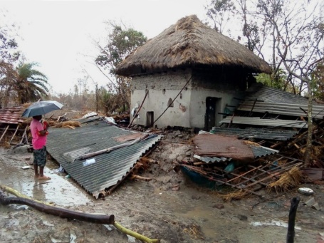 Ein Mann mit Regenschirm steht vor einem kleinen, gemauerten Haus, das von Trümmerteilen wie verbogenen Wellblechabdeckungen umgeben ist.