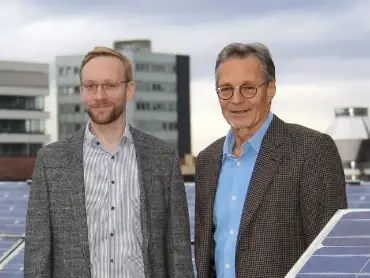 Auf dem Flachdach eines Hauses stehen ein jüngerer und ein etwas älterer Mann zwischen schräggestellten PV Modulen, dahinter Hochhäuser vor einem abendlichen Himmel..