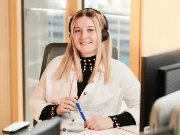 Eine freundllich lächelnde Frau mit Stift in der Hand und einem an einem Computer vor hellen Holzflächen und einem Fenster
