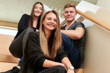 Drei junge Mitarbeitende sitzen auf einer Treppe in einem lichten Gebäude und lächeln in die Kamera.