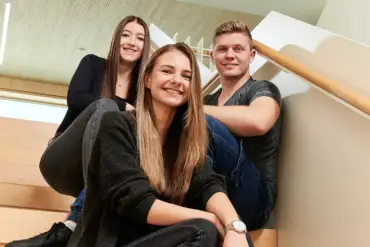 Dreii junge Mitarbeitende sitzen auf einer Treppe in einem lichten Gebäude und lächeln in die Kamera.