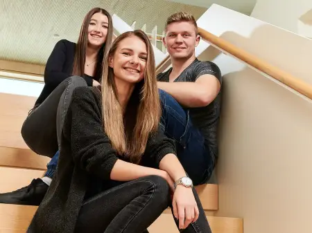 Drei junge Mitarbeitende sitzen auf einer Treppe in einem lichten Gebäude und lächeln in die Kamera.