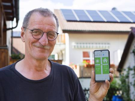Ein älterer Mann mit runder Brille und Schnauzbart hält in seiner linen Hand ein Smartphone mit einer Energie-App, im Hintergrund eine PV-Dachanlage