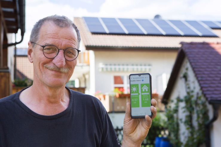 Ein älterer Mann mit runder Brille und Schnauzbart hält in seiner linen Hand ein Smartphone mit einer Energie-App, im Hintergrund eine PV-Dachanlage