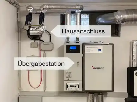 In einem Heizungskeller ein Nahwärmeanschluss und die reisekoffergroße Übergabestation, beide beschriftet.