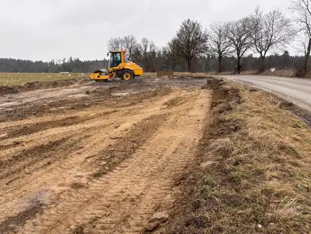 Ein gelber Bagger verrichtet Planierarbeiten auf einer Fläche neben einer Landstraße.