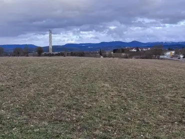 Eine brachliegende Landwirtschaftsfläche, im Hintergrund ein schneckenartig verkleideter Turm und bewaldete Höhenzüge