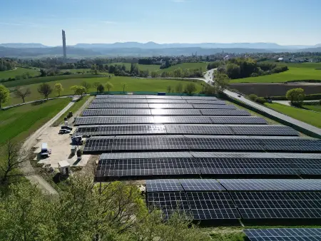 Ein Solarpark zwischen Wald und Wiesen, im Hintergrund ein schraubenartig verkleideter Turm und bewaldete Höhenzüge