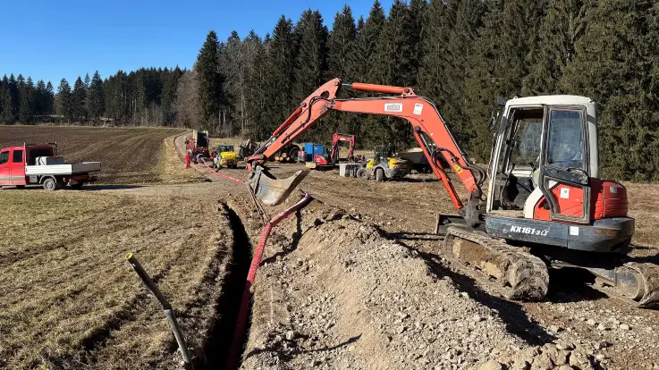 Im Vordergrund ein Graben, in dem eine Leitung verlegt wird, daneben und dahinter Bagger und weiteres schweres Baugerät, im Hintergrund ein Waldrand