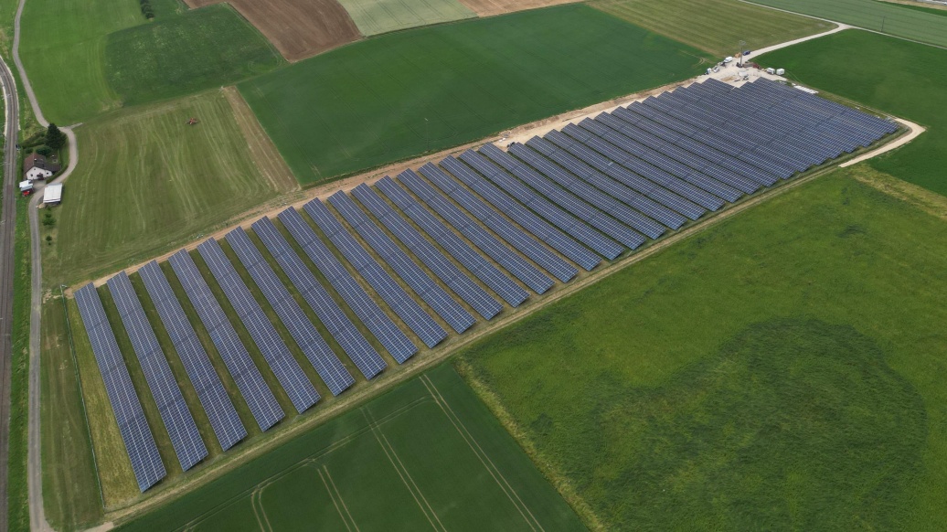 Blick auf ein Solarkraftwerk mit über 30 Modulreihen, im Hintergrund ein Dorf inmitten flach gewellter Landschaft