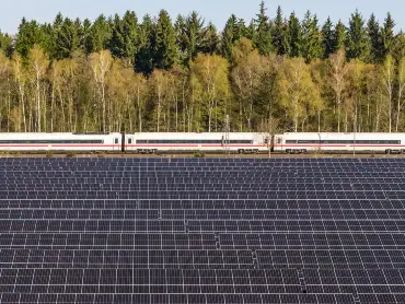 Ein Solarpark, dahinter fährt ein Zug auf der Bahnstrecke vor einem Wald.