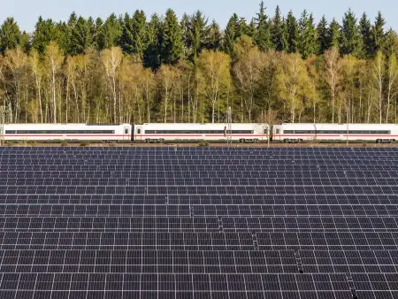 Ein Solarpark, dahinter fährt ein Zug auf der Bahnstrecke vor einem Wald.
