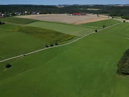 Luftaufname von Wiesen- und Feldflächen, dahinter sind ein Dorf und bewaldete Hügel zu erkennen