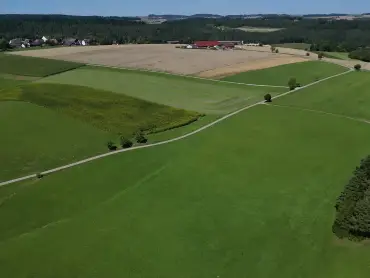 Luftaufname von Wiesen- und Feldflächen, dahinter sind ein Dorf und bewaldete Hügel zu erkennen