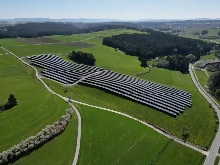 Luftaufname eines Solarparks zwischen Wiesen- und Feldflächen und einer Straße, dahinter ein Dorf und bewaldete Hügel.