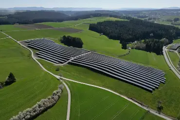 Luftaufname eines Solarparks zwischen Wiesen- und Feldflächen und einer Straße, dahinter ein Dorf und bewaldete Hügel.