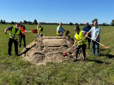 Ein Sandhaufen auf einem großen Feld, darum haben sich sieben Personen mit Spaten versammelt und zelebrieren den symbolischen Spatenstich.