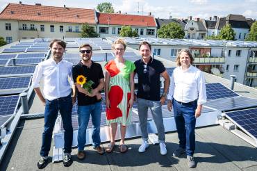 Fünf Menschen auf dem Flachdach eines Mehrfamilienhauses, hinter ihnen PV-Module
