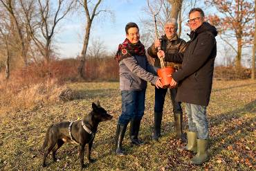 Auf einer Wiese stehen zwei Männer und eine Frau, die zu dritt einen Pflanztopf mit Baumsetzling halten, daneben ein Hund