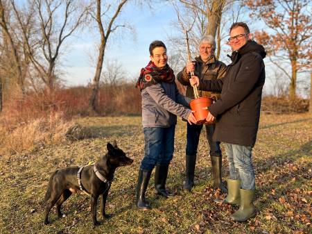 Auf einer Wiese stehen zwei Männer und eine Frau, die zu dritt einen Pflanztopf mit Baumsetzling halten, daneben ein Hund