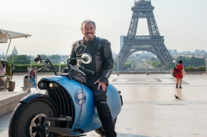 Ein E-Motorradfahrer posiert mit seiner Maschine auf einem großen Platz, im Hintergrund der Eiffelturm.