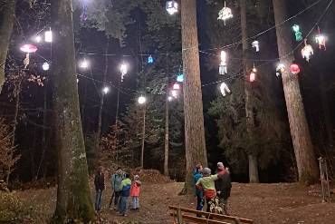 In einem nächtlichen Wald steht eine Gruppe von Menschen, über ihnen leuchten bunte Laternen an einer Kette