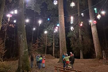In einem nächtlichen Wald steht eine Gruppe von Menschen, über ihnen leuchten bunte Laternen an einer Kette