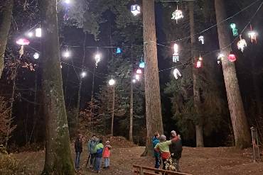 In einem nächtlichen Wald steht eine Gruppe von Menschen, über ihnen leuchten bunte Laternen an einer Kette