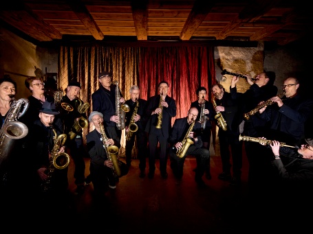 14 Menschen mit Saxofonen stehen im Halbkreis auf einer Theaterbühne