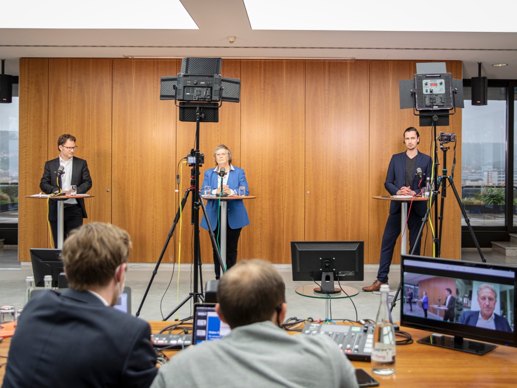 Blick in den Raum auf eine Podiumsdiskussion, vorne stehen eine Frau und zwei Männer, im Bildvordergrund zwei Veranstaltungstechniker mit einem Monitor.