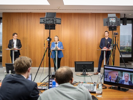 Blick in den Raum auf eine Podiumsdiskussion, vorne stehen eine Frau und zwei Männer, im Bildvordergrund zwei Veranstaltungstechniker mit einem Monitor.