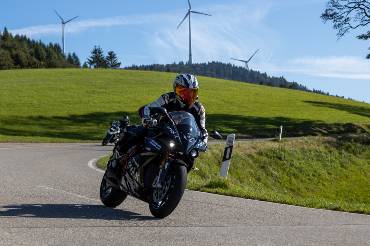 Ein E-Motorradfahrer legt sich in die Kurve, im Hintergrund ein Windrad