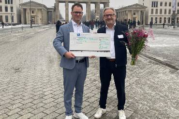 Zwei Männer stehen nebeneinander vor dem Brandenburger Tor, einer hält einen großen Blumenstrauß und eine Tafel, auf der ein Scheck abgebildet ist