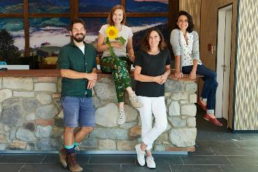 Drei Frauen und ein Mann sind an einem gemauerten Steintresen gruppiert, eine hält eine Sonnenblume.