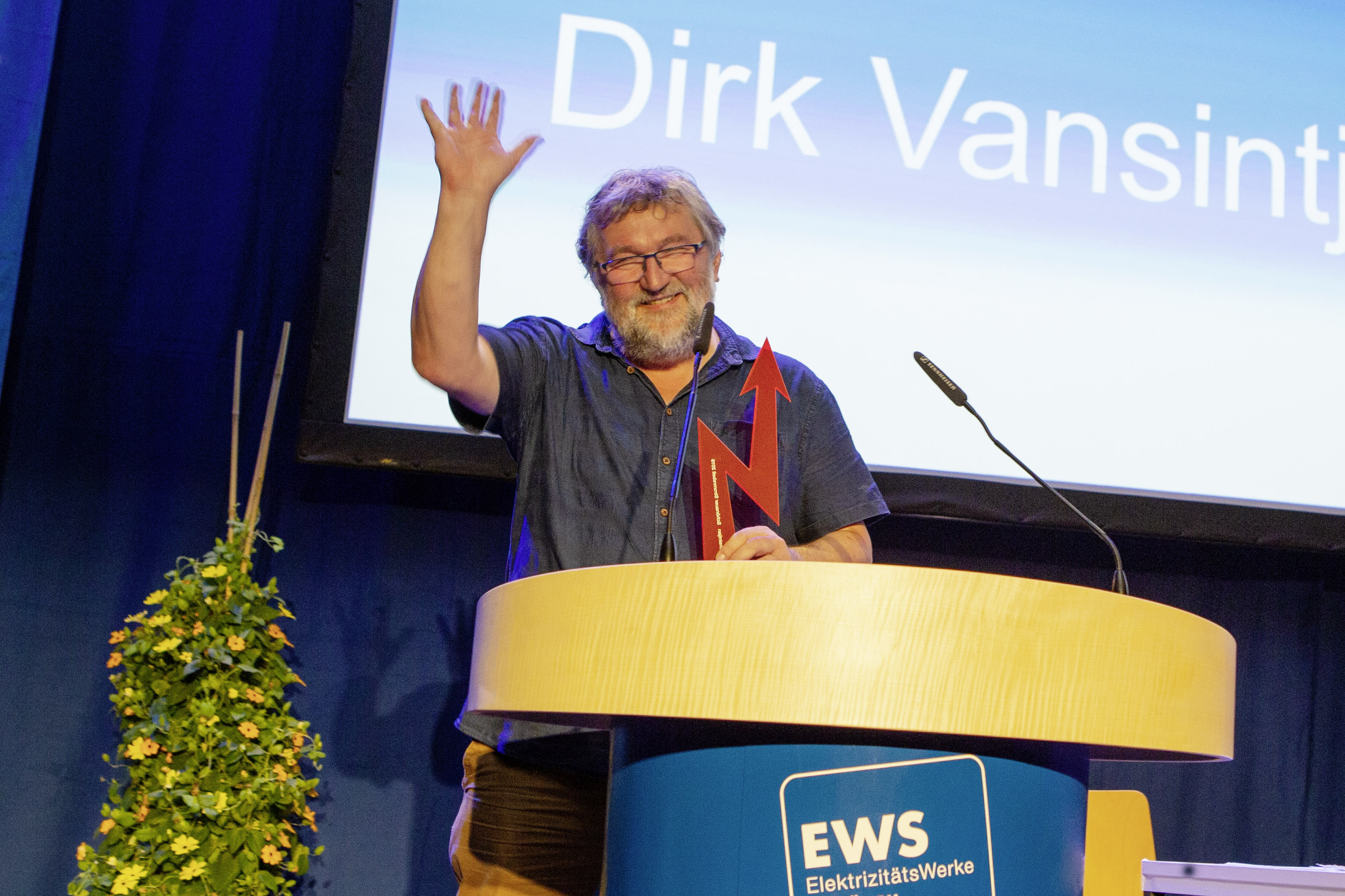 Dirk Vansintjan hebt – an einem Vortragspodium stehend – grüßend die Hand.