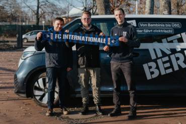 Drei Männer stehen vor einem VW-Elektrobus und halten zusammen einen Fanschal des FC Internationale Berlin hoch