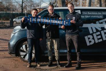 Drei Männer stehen vor einem VW-Elektrobus und halten zusammen einen Fanschal des FC Internationale Berlin hoch