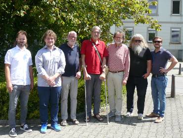 Gruppenfoto von sieben Männern nebeneinander auf der Straße vor einer Hecke