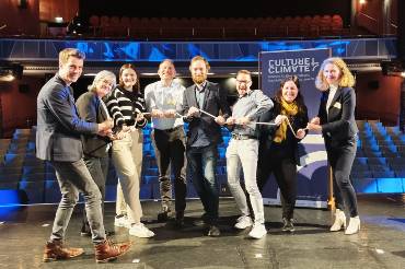 Acht Menschen stehen im Halbkreis auf einer Theaterbühne und halten gemeinsam ein Seil in den Händen.