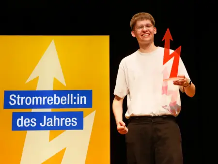 Ein lächelnder junger Mann mit Brille auf einer Bühne; in der Hand eine blitzförmige Trophäe, im Hintergrund ist auf einem Display «Stromrebell:in des Jahres» zu lesen.