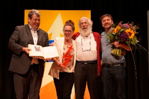 Auf einer Bühne stehen ein Mann mit einem Buch, eine Frau mit Trophäe in der Hand, ein älterer bärtiger Herr und ein Mann mit einem großen Blumenstrauß im Arm