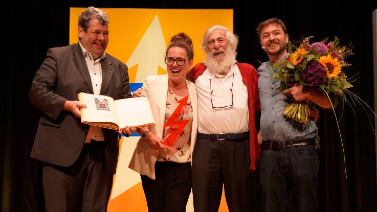 Auf einer Bühne stehen ein Mann mit einem Buch, eine Frau mit Trophäe in der Hand, ein älterer bärtiger Herr und ein Mann mit einem großen Blumenstrauß im Arm