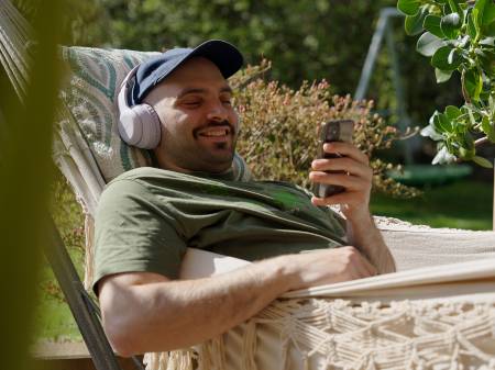 Ein junger Mann mit Kopfhörern liegt entspannt lächelnd in einer Hängematte im Freien; er schaut auf sein Smartphone.