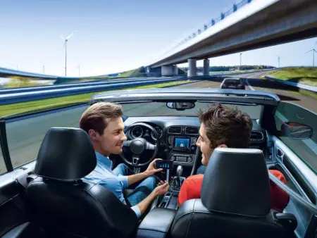 Zwei Männer sitzen in einem offenen Sportcoupé in voller Geschwindigkeit auf der Stadtautobahn, der Mann am Steuer zeigt seinem Beifahrer etwas an seinem Telefon und hat keine Hand am Steuer