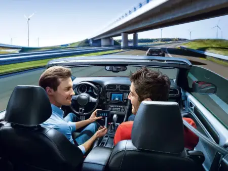 Zwei Männer sitzen in einem offenen Sportcoupé in voller Geschwindigkeit auf der Stadtautobahn, der Mann am Steuer zeigt seinem Beifahrer etwas an seinem Telefon und hat keine Hand am Steuer